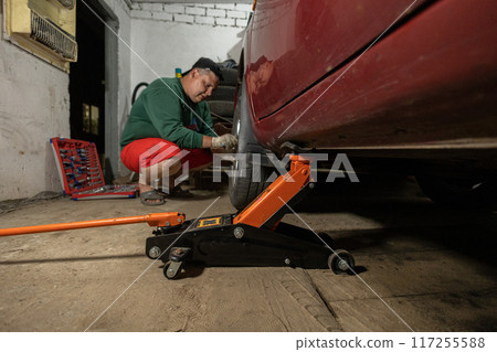 A car in a garage is raised on a jack while a man changes a tire. A man repairs a flat tire on a car in a garage. The concept of replacing a wheel on a car. 117255588
