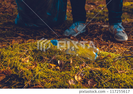 A plastic bottle lies on the moss in the sun. In the forest, a woman volunteer came up with a garbage bag to pick up plastic waste. Pollution of the forest with household waste and plastic garbage. A plastic bottle lies on the moss in the sun. In the forest, a woman volunteer came up with a garbage bag to pick up plastic waste. Pollution of the forest with household waste and plastic garbage. 117255617
