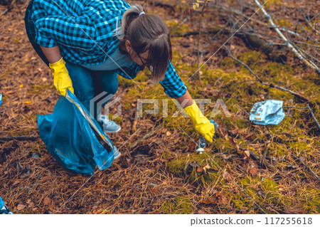 Saving the forest from garbage. A female volunteer collects plastic and glass waste from the forest into a garbage bag. A woman fights to save and preserve nature from garbage. Eco activism. Saving the forest from garbage. A female volunteer collects plastic and glass waste from the forest into a garbage bag. A woman fights to save and preserve nature from garbage. Eco activism. 117255618