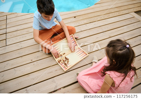 Children playing backgammon outdoors by the pool on a wooden deck Children playing backgammon outdoors by the pool on a wooden deck 117256175