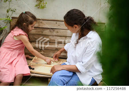 A mother and daughter spend quality time baking croissants together in a cozy kitchen. The little girl thoughtfully arranges baked goods while learning the art of cooking. 117256486