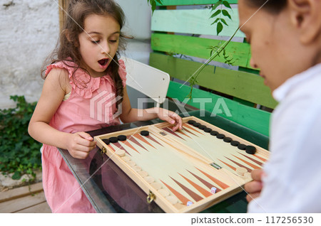 Young girl in pink dress playing an exciting game of backgammon with a friend outdoors Young girl in pink dress playing an exciting game of backgammon with a friend outdoors 117256530