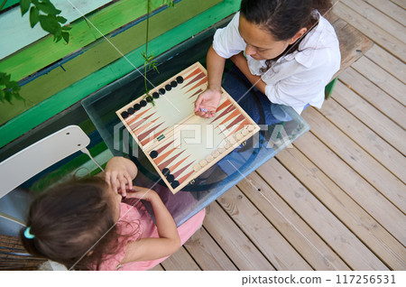 Mother and daughter playing backgammon at a table in an outdoor setting 117256531