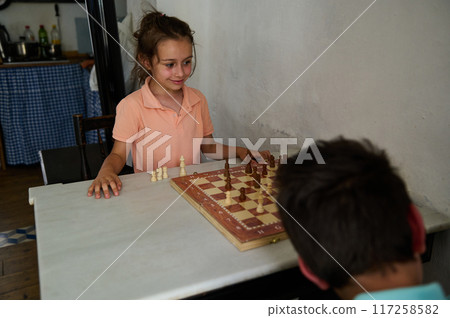 Young girl and boy playing chess indoors with focused expressions and casual clothing Young girl and boy playing chess indoors with focused expressions and casual clothing 117258582