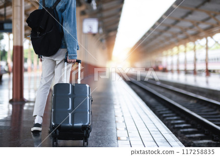 A young man holding a suitcase waits for a train at the train station for traveling. 117258835