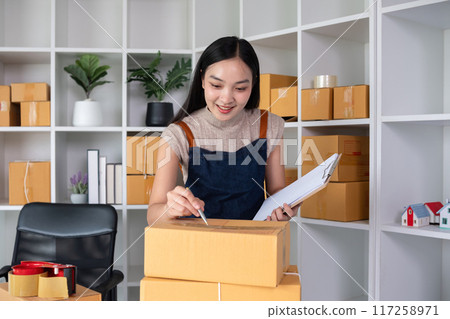 A small business owner receives product orders and writes shipping information on cardboard boxes in the home office to prepare for delivery. A small business owner receives product orders and writes shipping information on cardboard boxes in the home office to prepare for delivery. 117258971