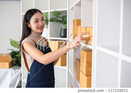 A small SME business owner checks an order and writes shipping information on a cardboard box in his home office to prepare for delivery. A small SME business owner checks an order and writes shipping information on a cardboard box in his home office to prepare for delivery. 117258972