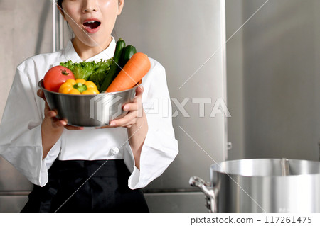A young female chef is surprised while holding a lot of vegetables A young female chef is surprised while holding a lot of vegetables 117261475