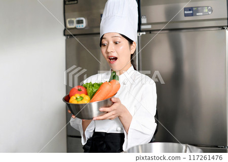 A young female chef is surprised while holding a lot of vegetables A young female chef is surprised while holding a lot of vegetables 117261476