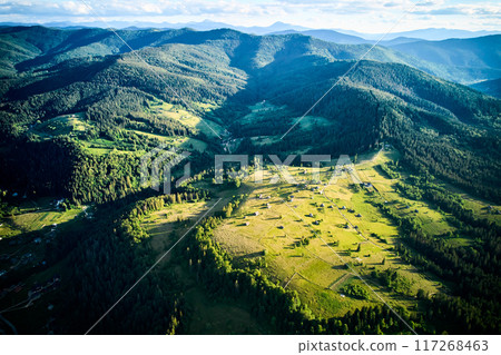 Aerial view of sunlit valley, dotted with small houses and winding roads, surrounded by dense forests. Golden light casts long shadows. Vibrant landscape stretches out, merging into forested hills. 117268463