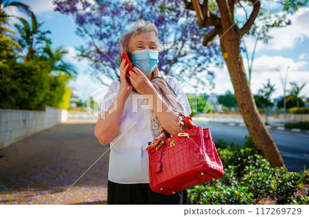 An 80-year-old woman, in a white blouse and face mask, smiles while talking on her red phone and holding a red handbag under a jacaranda tree on a city sidewalk. An 80-year-old woman, in a white blouse and face mask, smiles while talking on her red phone and holding a red handbag under a jacaranda tree on a city sidewalk. 117269729