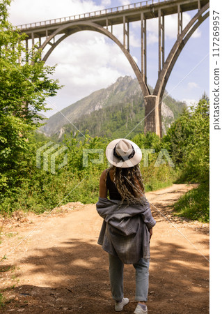 A girl in a hat stands against the backdrop of a beautiful arched bridge over the Tara River, a popular tourist attraction in Montenegro. 117269957