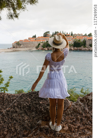 girl tourist in a hat looks from a top on the island of Sveti Stefan in Montenegro and the sea girl tourist in a hat looks from a top on the island of Sveti Stefan in Montenegro and the sea 117269958