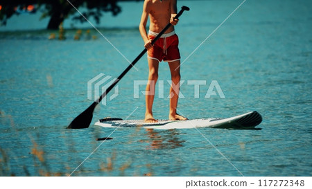 Unknown standing boy paddles a paddle board or SUP, summer vacation time Unknown standing boy paddles a paddle board or SUP, summer vacation time 117272348