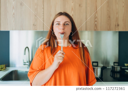 Portrait of excited hungry young woman holding fork in her mouth, biting and dreaming about delicious healthy food at the kitchen. Diet concept Portrait of excited hungry young woman holding fork in her mouth, biting and dreaming about delicious healthy food at the kitchen. Diet concept 117272361