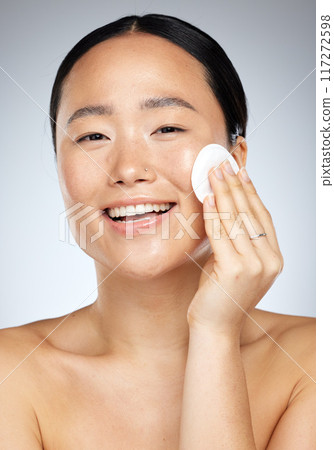 Skincare, beauty and woman with cotton pad for facial cleaning against a grey studio background. Happy, smile and portrait of a young Asian model with cotton for face wellness, clean body and makeup Skincare, beauty and woman with cotton pad for facial cleaning against a grey studio background. Happy, smile and portrait of a young Asian model with cotton for face wellness, clean body and makeup 117272598