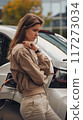 Woman stands next to white car with black roof and silver accents. Wears beige jacket and pants, contrasting with dark hair. Gray building in background. Charging electric car. 117273034