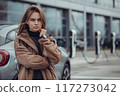 Young woman stands confidently on city street near electric car charging station. Wears stylish black jacket, beige trench coat. Modern building with large windows reflects surrounding environment. 117273042