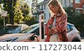 Young woman stands next to electric car charging. She wears pink jacket and white pants with serious expression. Charging station is gray, black shades with another car in background. 117273044
