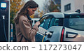 Woman stands near electric car at modern charging station on urban street. She holds black bag and looks at watch, waiting for car to charge. Electric vehicle concept photo with cityscape background. 117273047