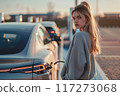 Woman stands beside electric car at modern charging station. Urban parking lot background. Gray sweatshirt, blue jeans worn by woman. Silver car with black roof rack. Electric Cars concept. 117273068