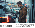 Man stands next to electric car at modern charging station. Looks at watch, waiting for charge. Electric vehicle concept, eco-friendly transportation, sustainable energy. 117273077