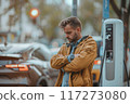 Man stands at electric car charging station, wearing brown jacket and blue jeans. Gazes off to side, urban backdrop with moving vehicles for modern transportation and eco-friendly lifestyle. 117273080