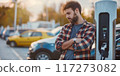 Man stands next to electric car charging station with various cars nearby. Wears blue shirt, arms crossed in relaxed manner. Charging car at modern eco-friendly station. 117273082