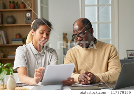 Portrait of young woman and senior man discussing project details in a modern home office, surrounded by books and office supplies, with light streaming through large windows Portrait of young woman and senior man discussing project details in a modern home office, surrounded by books and office supplies, with light streaming through large windows 117279199