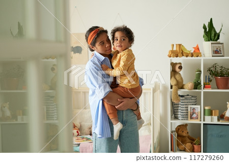 Woman lovingly holding her child in a cozy, well-decorated living space surrounded by toys and bookshelves, featuring plants and stuffed animals on shelves 117279260