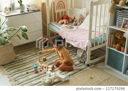 Young child playing with colorful blocks on carpeted floor in cozy bedroom filled with toys and stuffed animals, near crib and dresser Young child playing with colorful blocks on carpeted floor in cozy bedroom filled with toys and stuffed animals, near crib and dresser 117279266