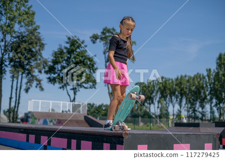 A girl in a bright pink skirt with a skateboard before a stunt at the grind boxes of the skatepark 117279425