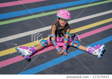 A beautiful girl in full protective gear and roller skates is sitting on the striped bright asphalt of the skatepark 117279431