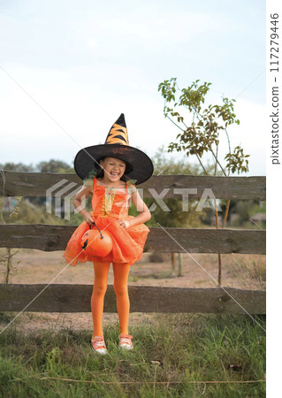 A laughing mischievous girl in a witch costume with a bucket in the shape of a pumpkin stands by the fence 117279446