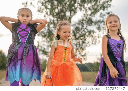 Funny girls in carnival costumes of witches and pumpkins walk down the street on Halloween eve 117279447