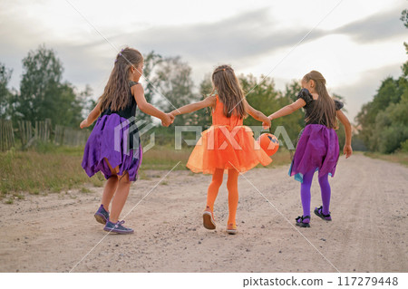 Girls in Halloween costumes run down a country street with a pumpkin-shaped bucket to collect festive sweets Girls in Halloween costumes run down a country street with a pumpkin-shaped bucket to collect festive sweets 117279448