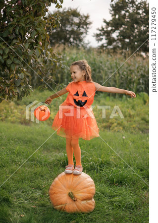 A girl in an orange costume stands on a large pumpkin with a bucket for collecting Halloween candies A girl in an orange costume stands on a large pumpkin with a bucket for collecting Halloween candies 117279450