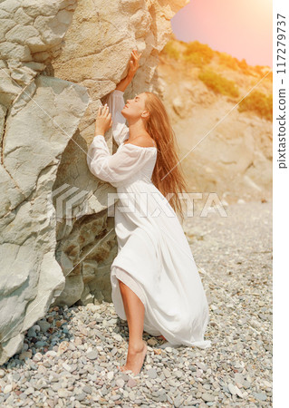 A woman in a white dress is standing on a rocky beach. Concept of serenity and tranquility, as the woman is enjoying the natural beauty of the beach. 117279737