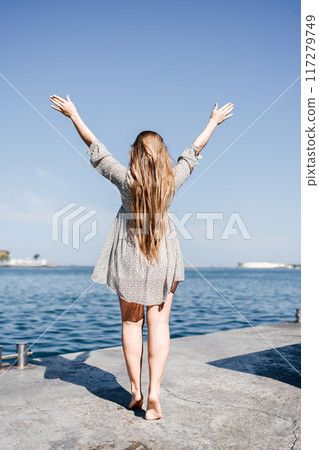 woman in a dress stands on a dock by the water, her arms raised in the air. Concept of freedom and joy, as the woman is celebrating or expressing her happiness. woman in a dress stands on a dock by the water, her arms raised in the air. Concept of freedom and joy, as the woman is celebrating or expressing her happiness. 117279749