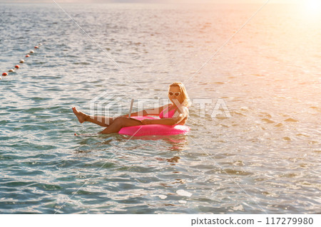 Woman works on laptop in sea. Freelancer, blond woman in sunglases floating on an inflatable big pink donut with a laptop in the sea at sunset. People summer vacation rest lifestyle concept. 117279980