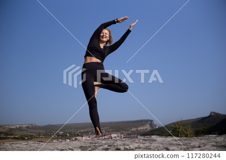 A woman is doing yoga on a rock with a blue sky in the background 117280244