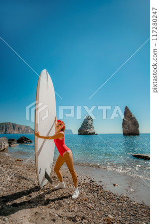Woman surfer during summer travel to beach vacation. A middle-aged woman in a red swimsuit and a cap with sap on the background of the sea and rocks in the water. extreme sport. Travel, weekend 117280247