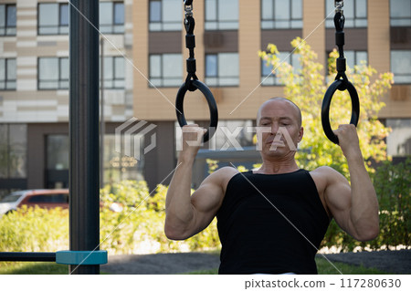 Athlete muscular fitness male pulling up on gymnastic rings at street gym. Outdoors. Summer. Workout routine. Athlete muscular fitness male pulling up on gymnastic rings at street gym. Outdoors. Summer. Workout routine. 117280630