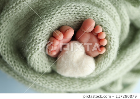The tiny foot of a newborn baby. Soft feet of a new born in a olive green wool blanket. Close up of toes, heels and feet of a newborn. Knitted white heart in the legs of a baby. Macro photography. The tiny foot of a newborn baby. Soft feet of a new born in a olive green wool blanket. Close up of toes, heels and feet of a newborn. Knitted white heart in the legs of a baby. Macro photography. 117280879