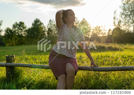 A woman relaxes on a fence while soaking in the sunset A woman relaxes on a fence while soaking in the sunset 117281900