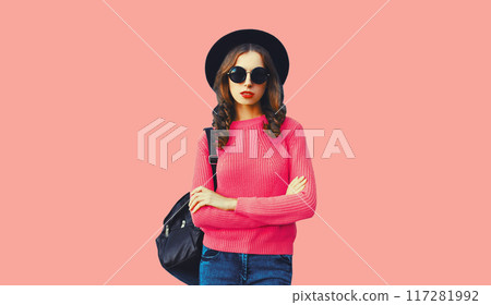 Modern young woman posing wearing sweater, black round hat and backpack on pink studio background 117281992