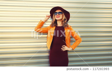 Beautiful smiling young woman wearing orange jacket, round hat on gray background Beautiful smiling young woman wearing orange jacket, round hat on gray background 117281993