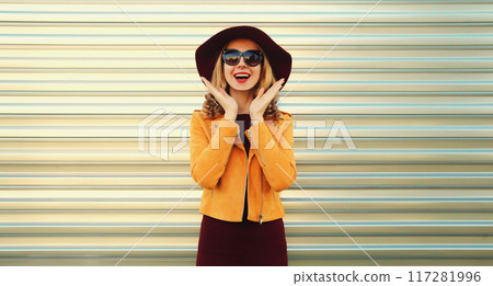 Happy excited surprised young woman wearing orange jacket, round hat on gray background 117281996
