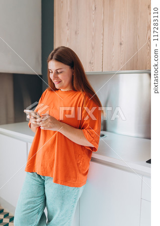 A happy 30s woman standing in a kitchen, holding a cell phone at home. Domestic life and leisure moments. High quality photo. 117283110