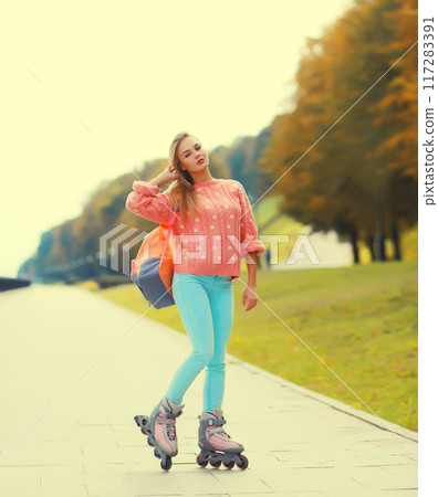 Stylish blonde young woman on roller skates with backpack posing in city park 117283391